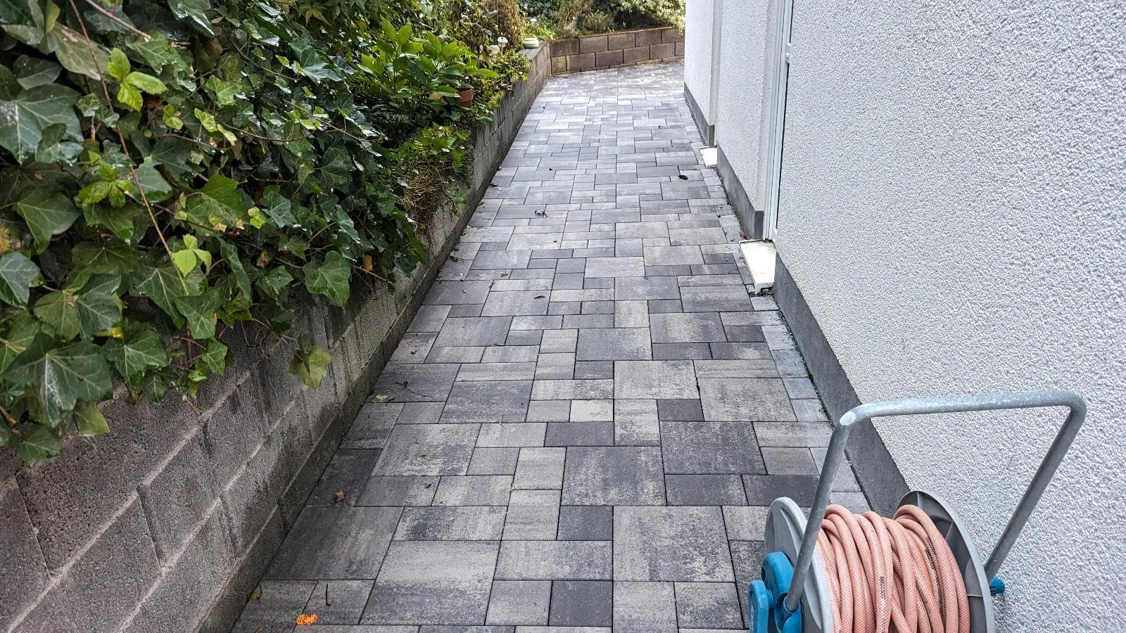 Schmaler Pflasterweg im Grauverbund entlang einer Hauswand mit Mauerkante